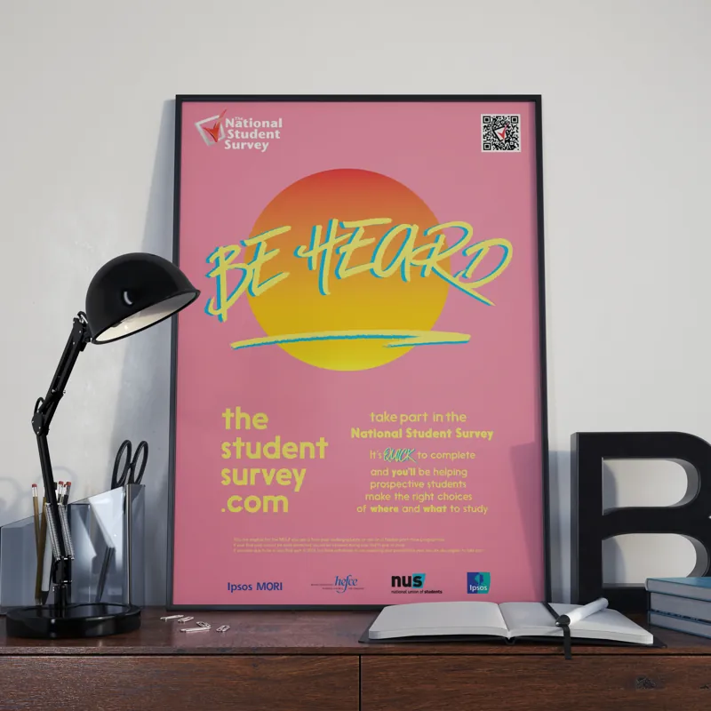 A framed poster on a desk: Be Heard in green script over an orange sun on pink, with the survey's address below.